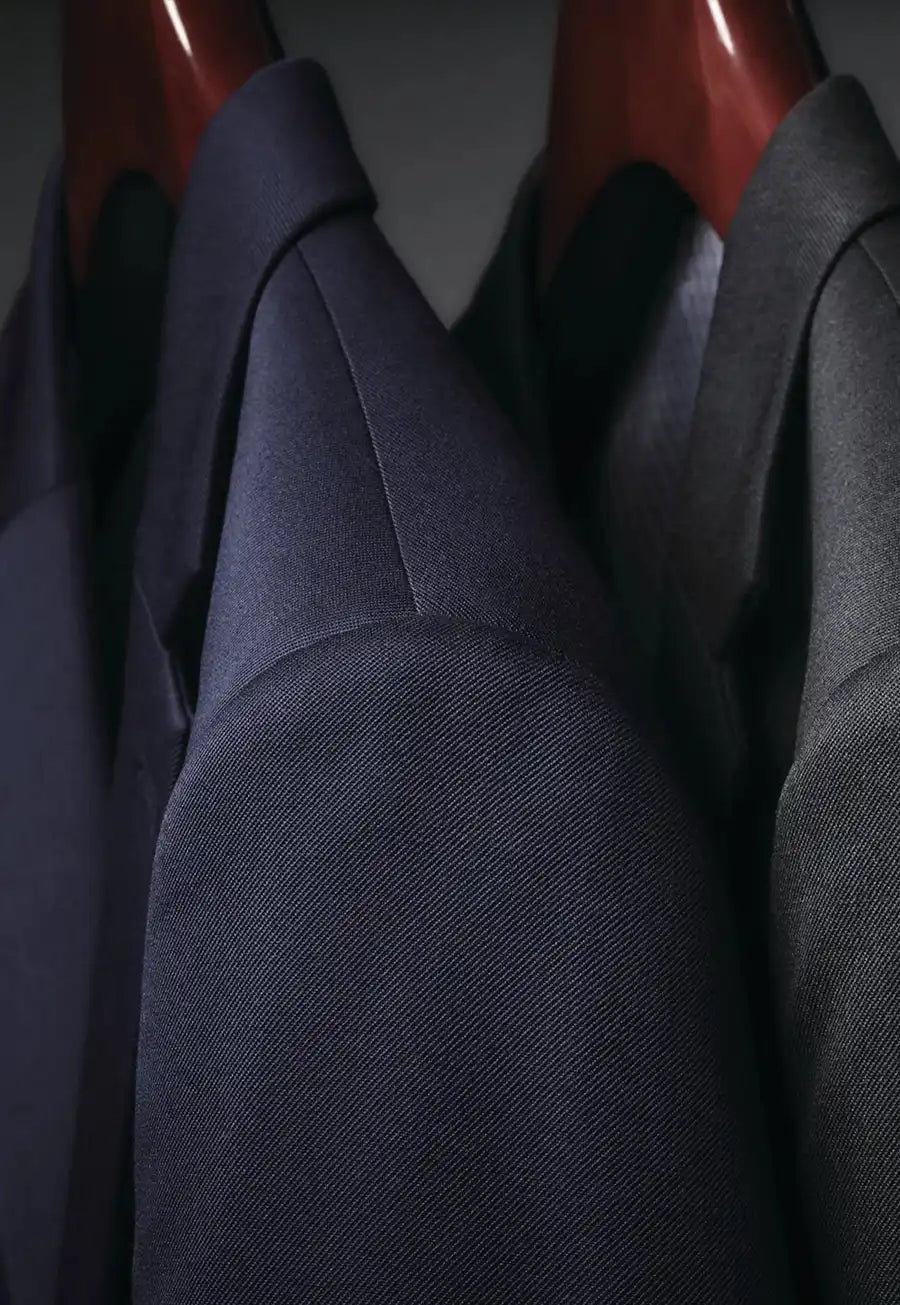 Close-up of several tailored Brook Taverner suit jackets from the Concept Collection hanging on wooden hangers, with the central jacket in dark navy blue and the surrounding jackets in charcoal gray and black.