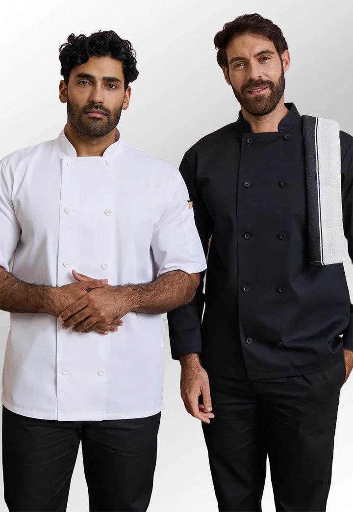 Two men modeling chef jackets against a plain light gray studio background. The man on the left wears a white short-sleeve chef jacket with black pants, and the man on the right wears a black double-breasted chef jacket with black pants and a white-and-black shoulder strap.