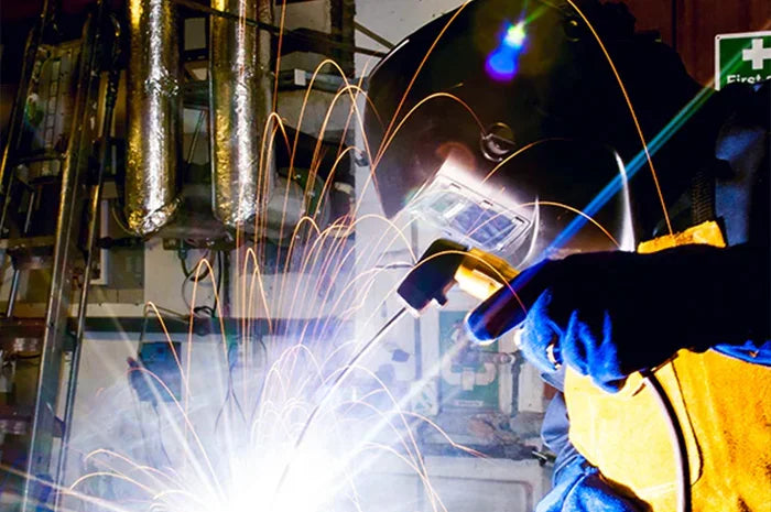 A welder in a black helmet and protective gloves holds a welding torch, producing bright sparks in an industrial setting with metal pipes in the background.