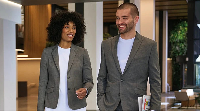 A man and a woman walking side-by-side in a modern office lobby. The woman is wearing a gray blazer over a white top, and the man is wearing a gray blazer over a white t-shirt.
