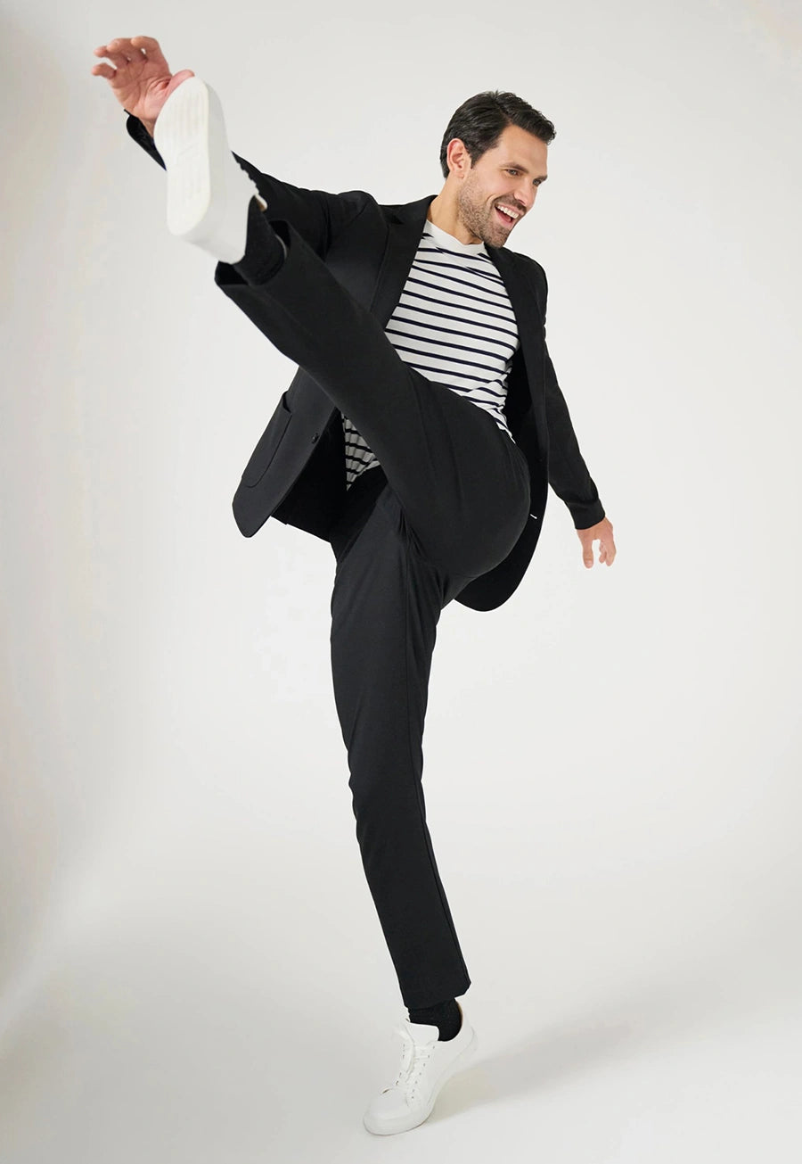 Model wearing the Brook Taverner Taurus Jersey Stretch Men's Trousers in Black poses dynamically with one leg raised high. 
