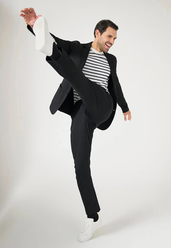 Model wearing the Brook Taverner Taurus Jersey Stretch Men's Trousers in Black poses dynamically with one leg raised high. 